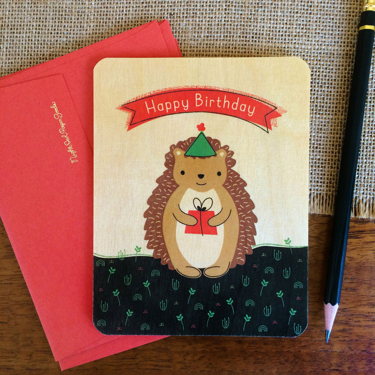 Happy Birthday card with cute hedgehog with green party hat holding a gift on birch wood with envelope