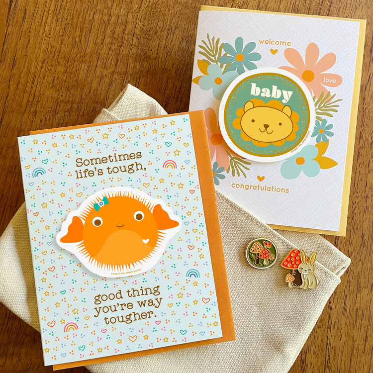 Assortment of new products, including Puffer Fish Sticker Encouragement Card, Baby Lion Sticker Congratulations and Mushrooms and Rabbit enamel pins 