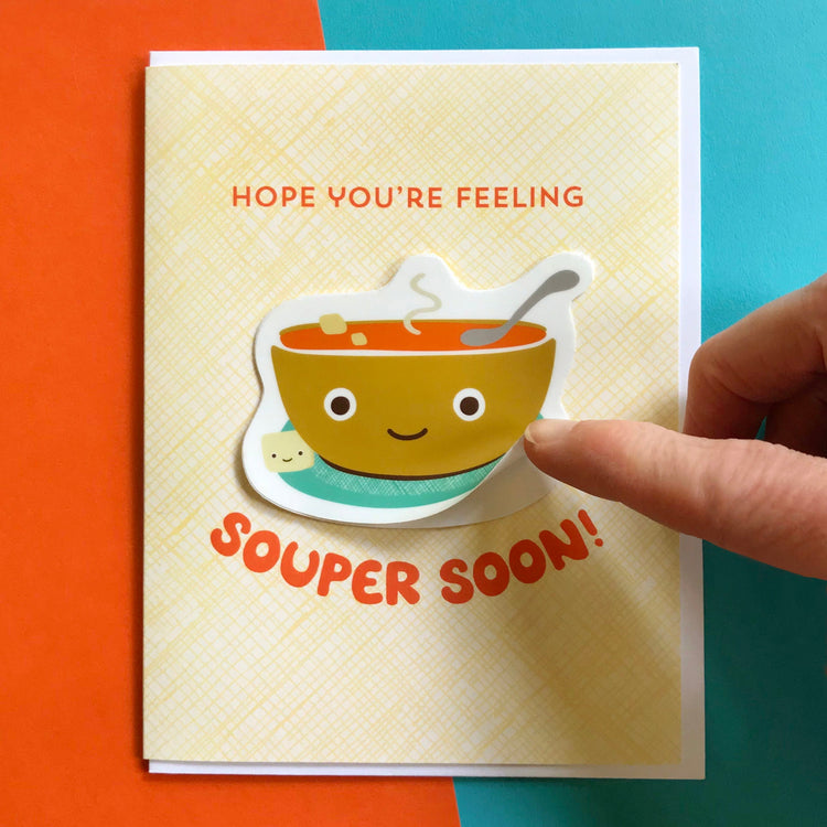 Get Well greeting card with cup of steaming tomato soup with happy, cute smiling face and happy cracker removable vinyl sticker. Finger is showing how the sticker can be removed from the card.