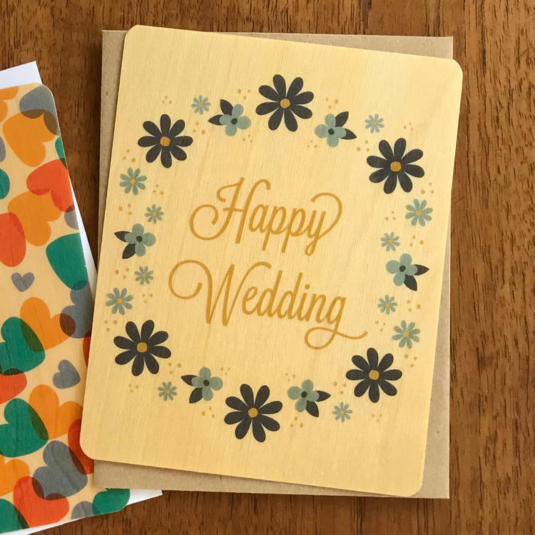 Light color birch wood flat wedding card with a circle of neutral blue, gray and navy flowers with the words 'Happy Wedding' in a golden color.