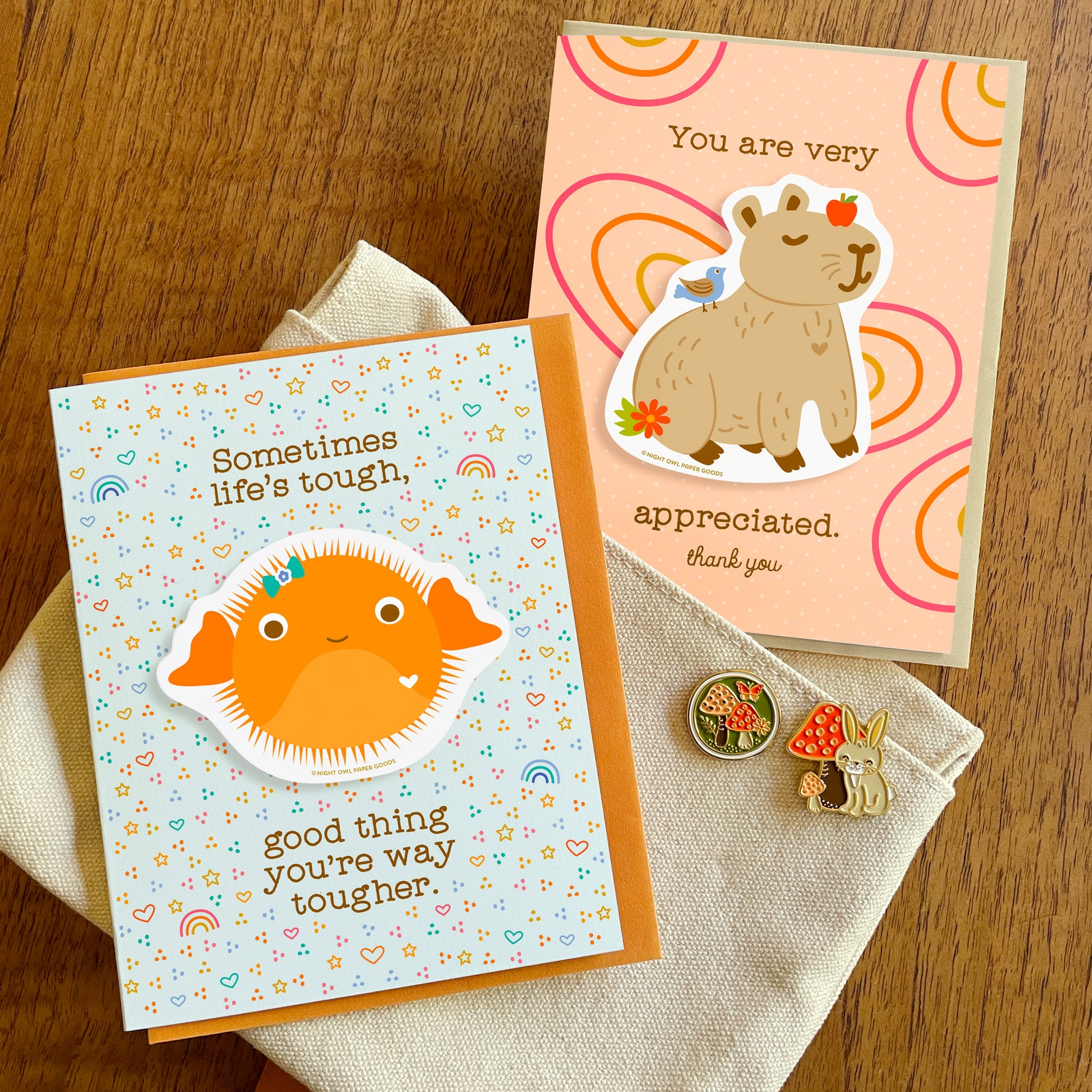 Encouragement card with a removable orange puffer fish vinyl sticker and the message “Sometimes life’s tough, good thing you’re way tougher” alongside a Thank You card with a removable vinyl capybara sticker and “You are very appreciated. Thank You” text. Also shown are a mushrooms enamel pin and a rabbit enamel pin.