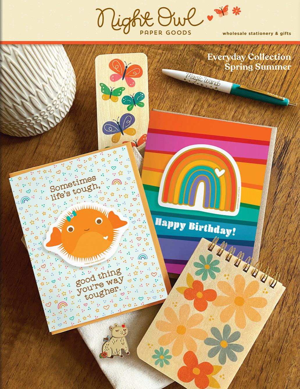Spring/Summer Wholesale Catalog Cover featuring a Puffer Fish sticker encouragement card, a Rainbow Stripes sticker birthday card, a butterfly wood bookmark, a friendly flower wood mini notepad a Capybara enamel pin and a Magic Wand ballpoint pen.