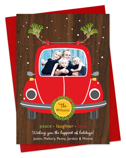 Customizable paper holiday card featuring your photo in the windshield of a red car with a printed walnut wood grain background. Features family name on the license plate and a personal greeting at the bottom of the card. Shown on a red envelope.
