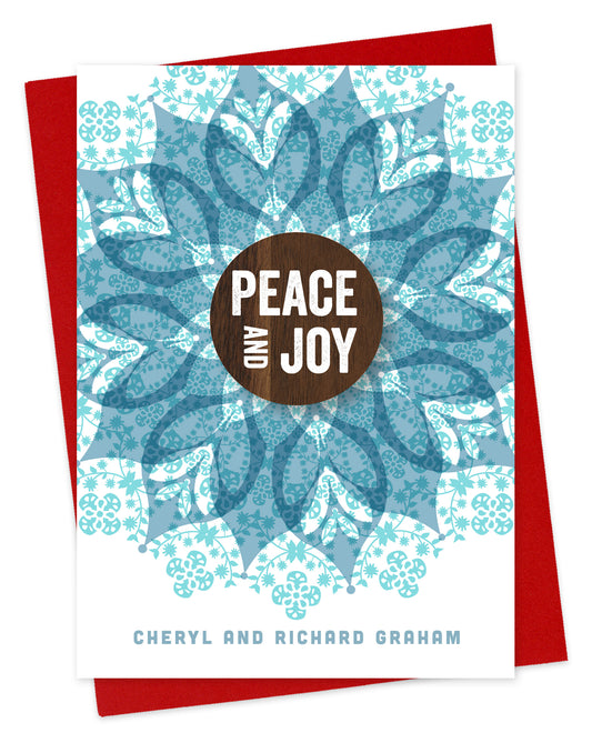 Customizable paper holiday card featuring a detailed blue blossom with a walnut wood grain medallion in the middle that reds “Peace and Joy.” Personal text is at the bottom. Shown with a red envelope.