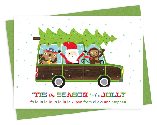 Front of customizable paper holiday card featuring Santa, a monkey and a beaver driving a wood-paneled station wagon with a chopped-down Christmas tree on top, “‘Tis the Season to be Jolly” text and a personal greeting. Shown on a green envelope.
