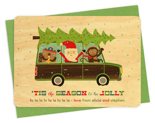 Front of customizable wood holiday card featuring Santa, a monkey and a beaver driving a wood-paneled station wagon with a chopped-down Christmas tree on top, “‘Tis the Season to be Jolly” text and a personal greeting. Shown on a green envelope.