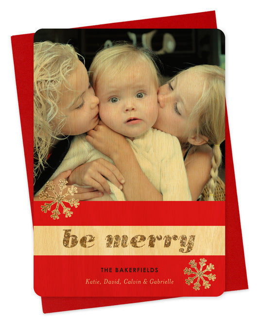 Customizable wood holiday card featuring a photo across the top, a red and birch wood striped banner with snowflakes and “Be Merry” in glittery gold font, plus a personal greeting. Shown with a red envelope.