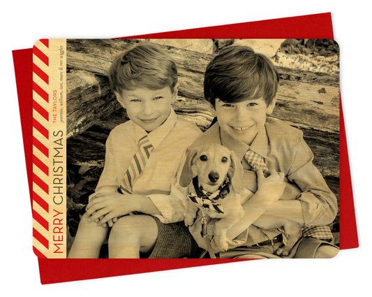 Customizable wood holiday card featuring a landscape photo with bold diagonal red stripes, “MERRY CHRISTMAS” text and a personal greeting running vertically along the left side. Shown with a red envelope.
