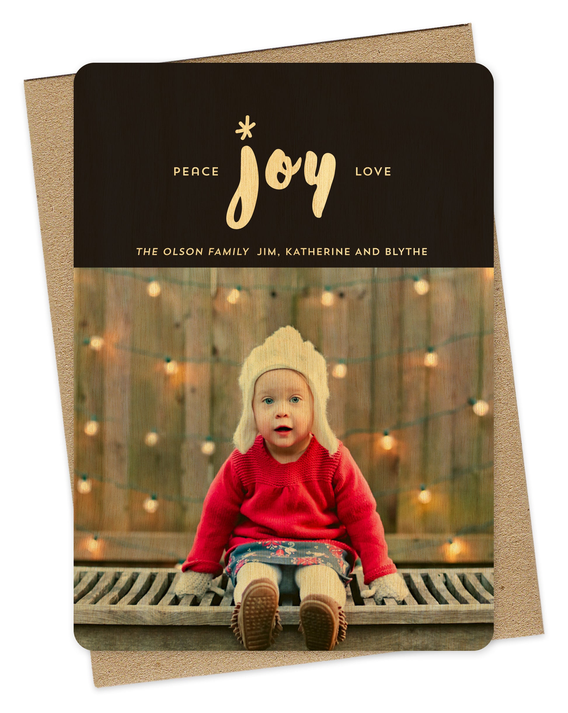 Customizable wood holiday card featuring your photo beneath a striking black banner adorned with “Peace Joy Love” text and a personal greeting. Shown with a kraft envelope.
