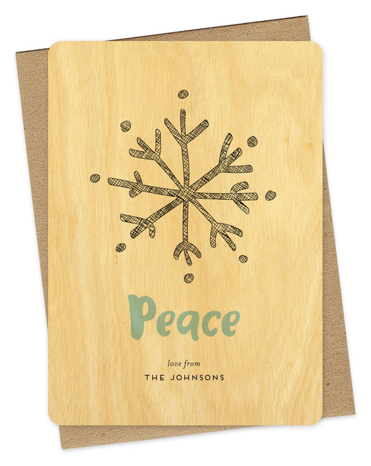 Customizable wood holiday card featuring a large illustrated snowflake, aqua "Peace” text and a personal greeting against a birch wood backdrop. Shown with a kraft envelope.