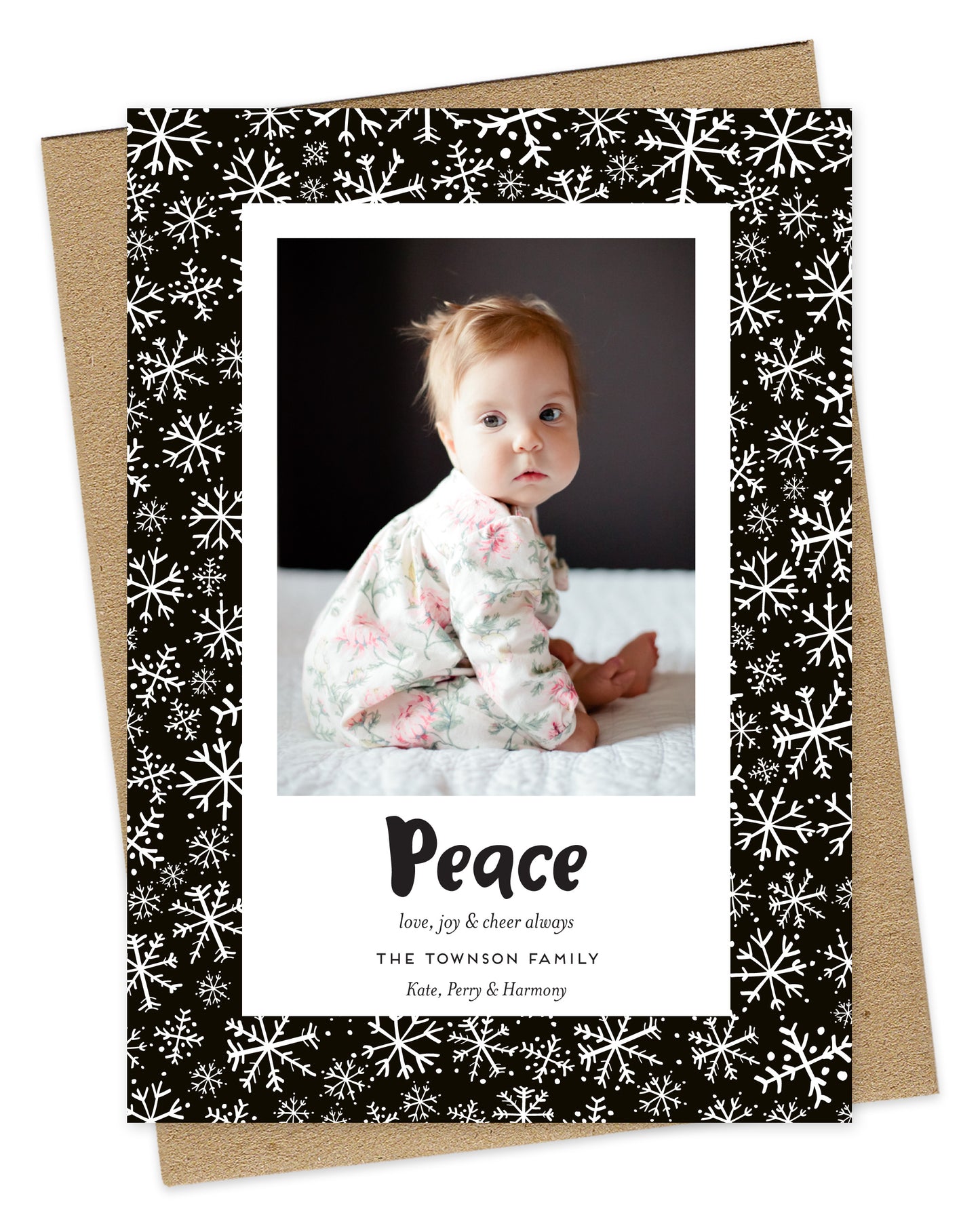 Customizable paper holiday card featuring a family photo with “Peace” text and a personal greeting near the bottom, framed by a black border with a snowflake pattern. Shown with a kraft envelope.