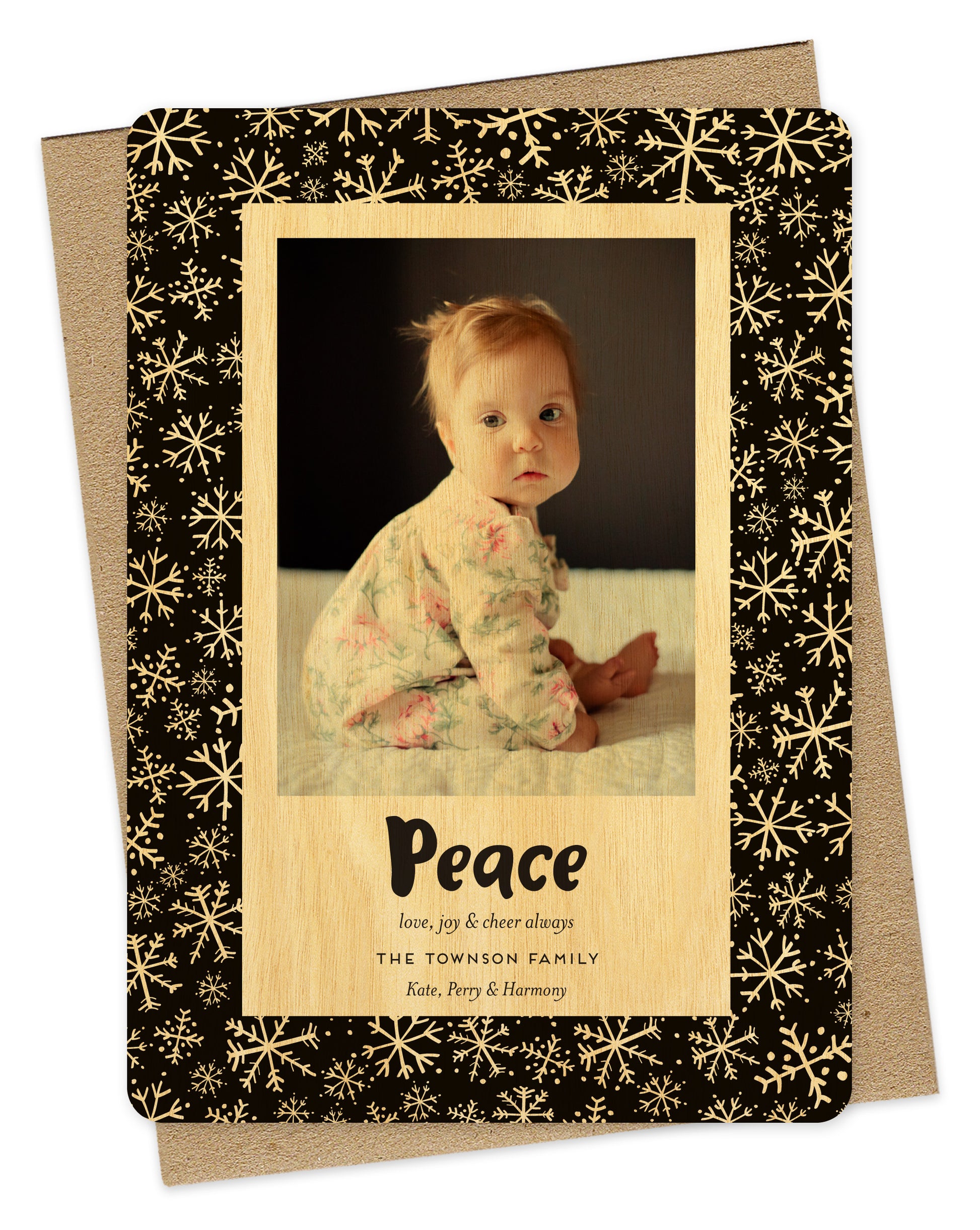 Customizable wood holiday card featuring a family photo with “Peace” text and a personal greeting near the bottom, framed by a black border with a snowflake pattern. Shown with a kraft envelope.