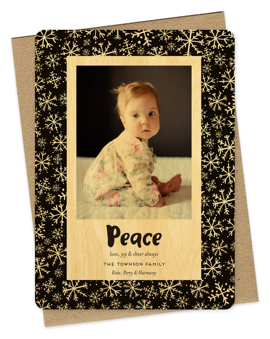 Customizable wood holiday card featuring a family photo with “Peace” text and a personal greeting near the bottom, framed by a black border with a snowflake pattern. Shown with a kraft envelope.