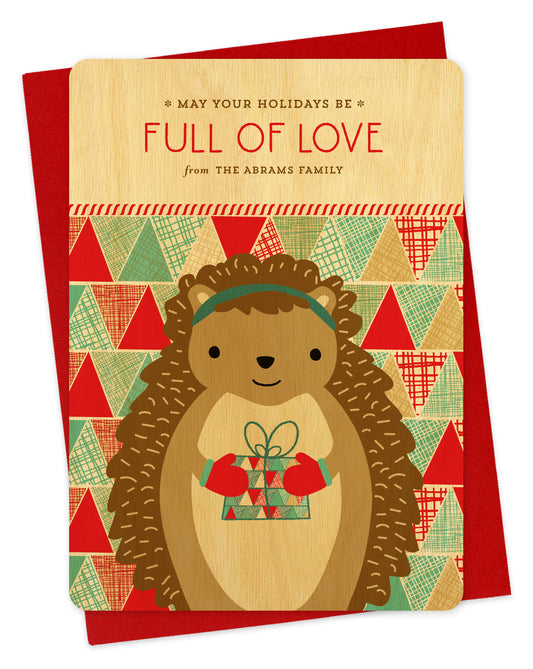 Customizable birch wood holiday card featuring a cute gift-giving hedgehog, “May Your Holidays Be Full Of Love” text and a personal greeting on a colorful, patchwork background. Shown with a red envelope.