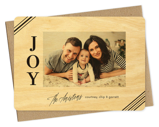 Customizable wood holiday card with a photo framed by a birch wood grain border, corner stripes, the word “JOY” on the left and a personal greeting on the bottom, all printed in black ink. Shown with a kraft envelope.