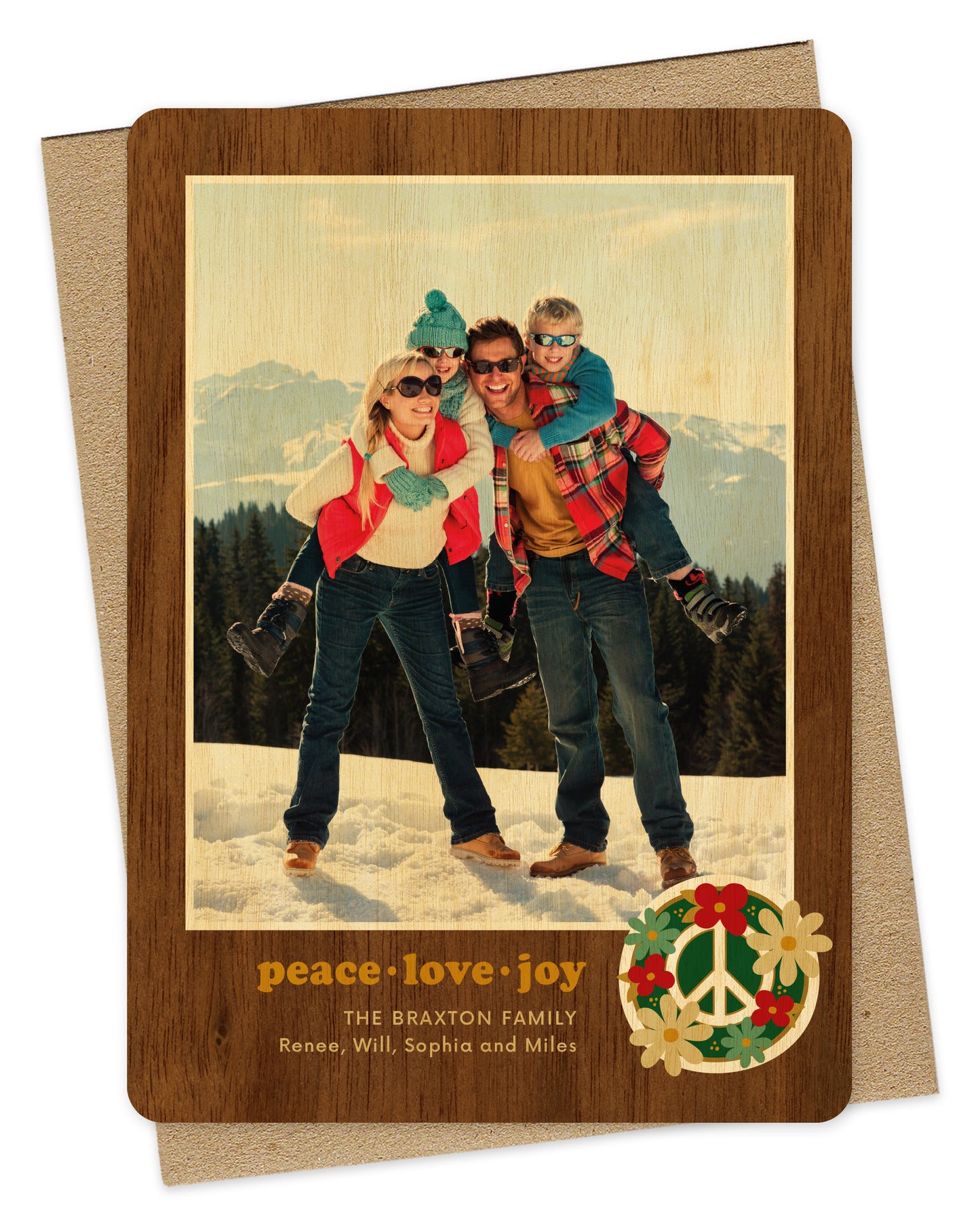 Customizable wood holiday card featuring a family photo with a printed walnut wood grain border and “peace love joy” text and a peace sign surrounded by flowers near the bottom. Shown with a kraft envelope.