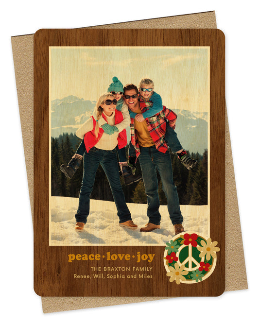 Customizable wood holiday card featuring a family photo with a printed walnut wood grain border and “peace love joy” text and a peace sign surrounded by flowers near the bottom. Shown with a kraft envelope.