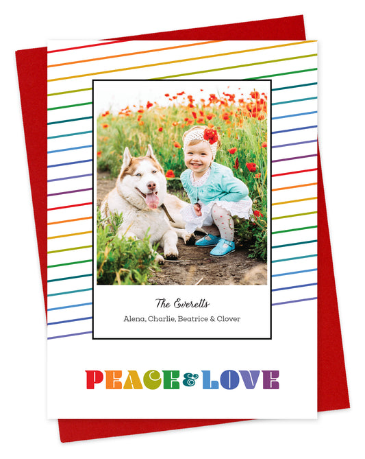Customizable paper holiday card featuring a photo bordered by thin, diagonal rainbow stripes and rainbow colored “PEACE & LOVE” text near the bottom, shown with a red envelope.
