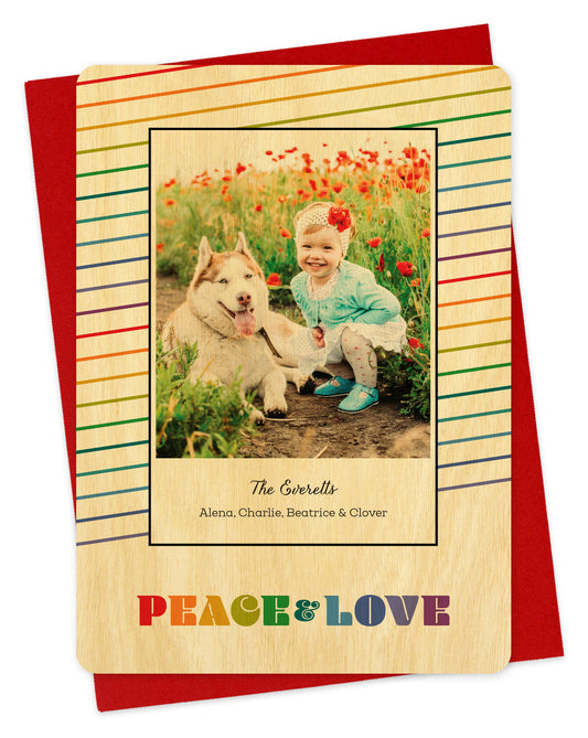 Customizable wood holiday card featuring a photo bordered by thin, diagonal rainbow stripes and rainbow colored “PEACE & LOVE” text near the bottom, shown with a red envelope.