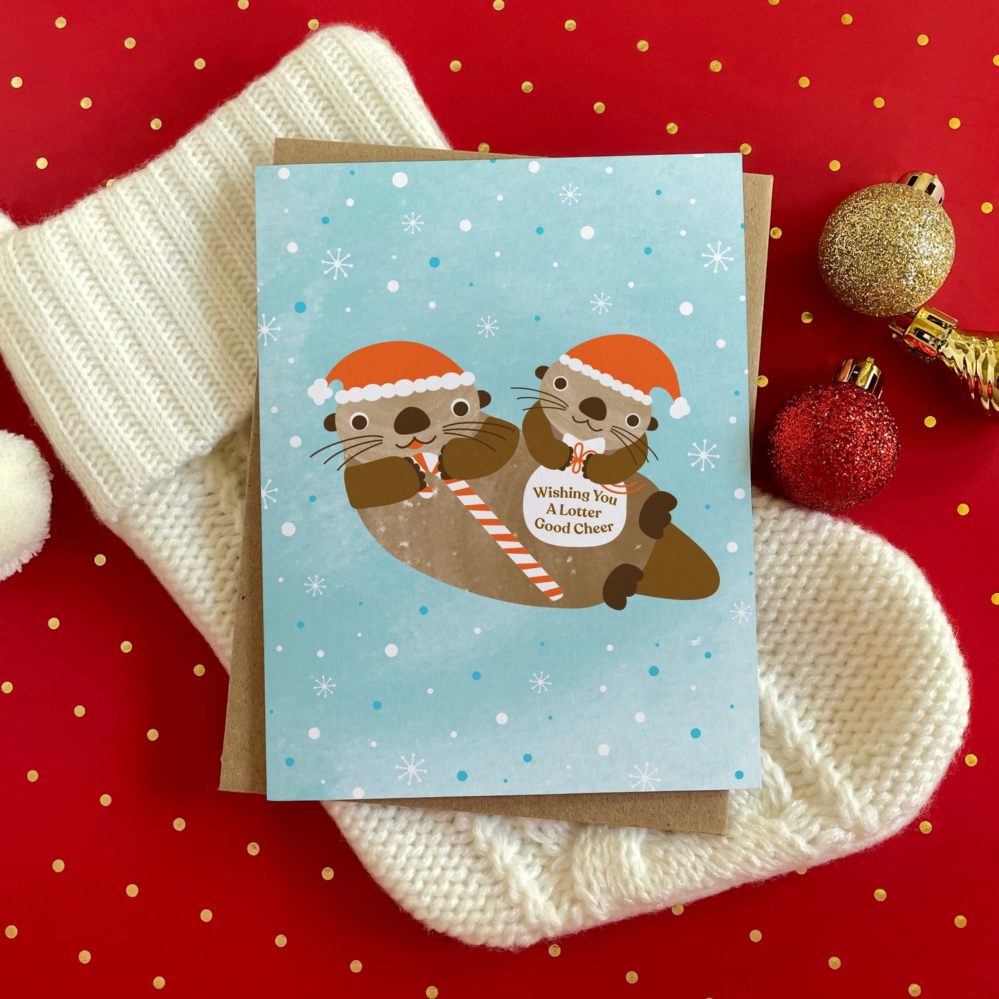 Folded Christmas card with a cute illustrated otter family wearing santa hats. One otter is holding a candy cane and the other is holding a bag that says 'Wishing You A Lotter Good Cheer.' Background of card is light blue with snowflakes. Card is sitting on top of a stocking with glittery red and gold ornaments nearby.
