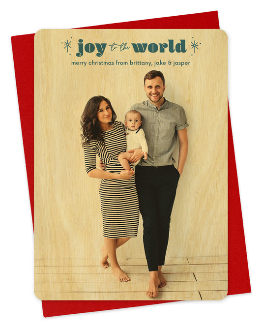 Customizable wood holiday card featuring a full portrait photo with "joy to the world” text and sparkle illustrations near the top, shown with a red envelope.