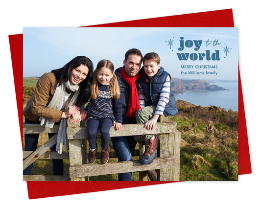 Customizable paper holiday card featuring a full landscape photo with "joy to the world” text, sparkle illustrations and a personal greeting near the top. Shown with a red envelope.