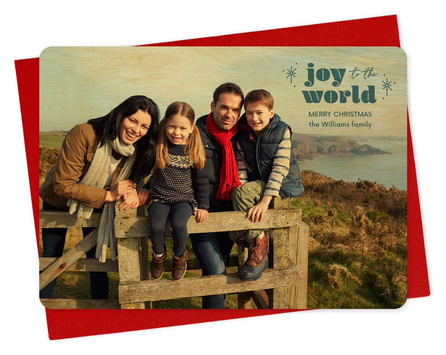 Customizable wood holiday card featuring a full landscape photo with "joy to the world” text, sparkle illustrations and a personal greeting near the top. Shown with a red envelope.