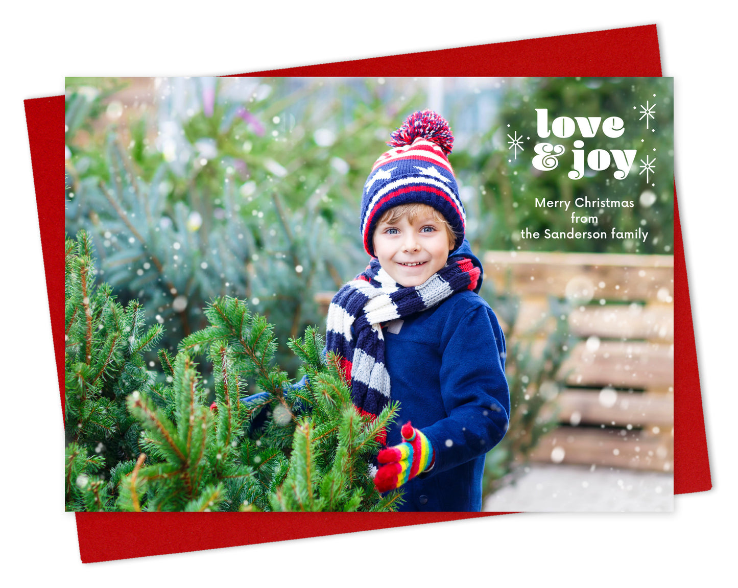 Customizable paper holiday card featuring a full landscape photo with "love & joy” text and sparkle illustrations near the top, shown with a red envelope.