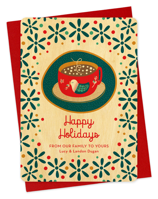 Front of wood holiday card featuring a mug of hot chocolate with a folk-style bird design, “Happy Holidays” text and a  personal greeting on a festive patterned background. Shown with a red envelope.