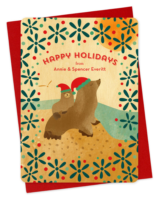 Customizable birch wood holiday card featuring two illustrated sea lions in Santa hats, near the water with a festive patterned background. “Happy Holidays” sentiment along with a personal greeting. Shown with a red envelope.