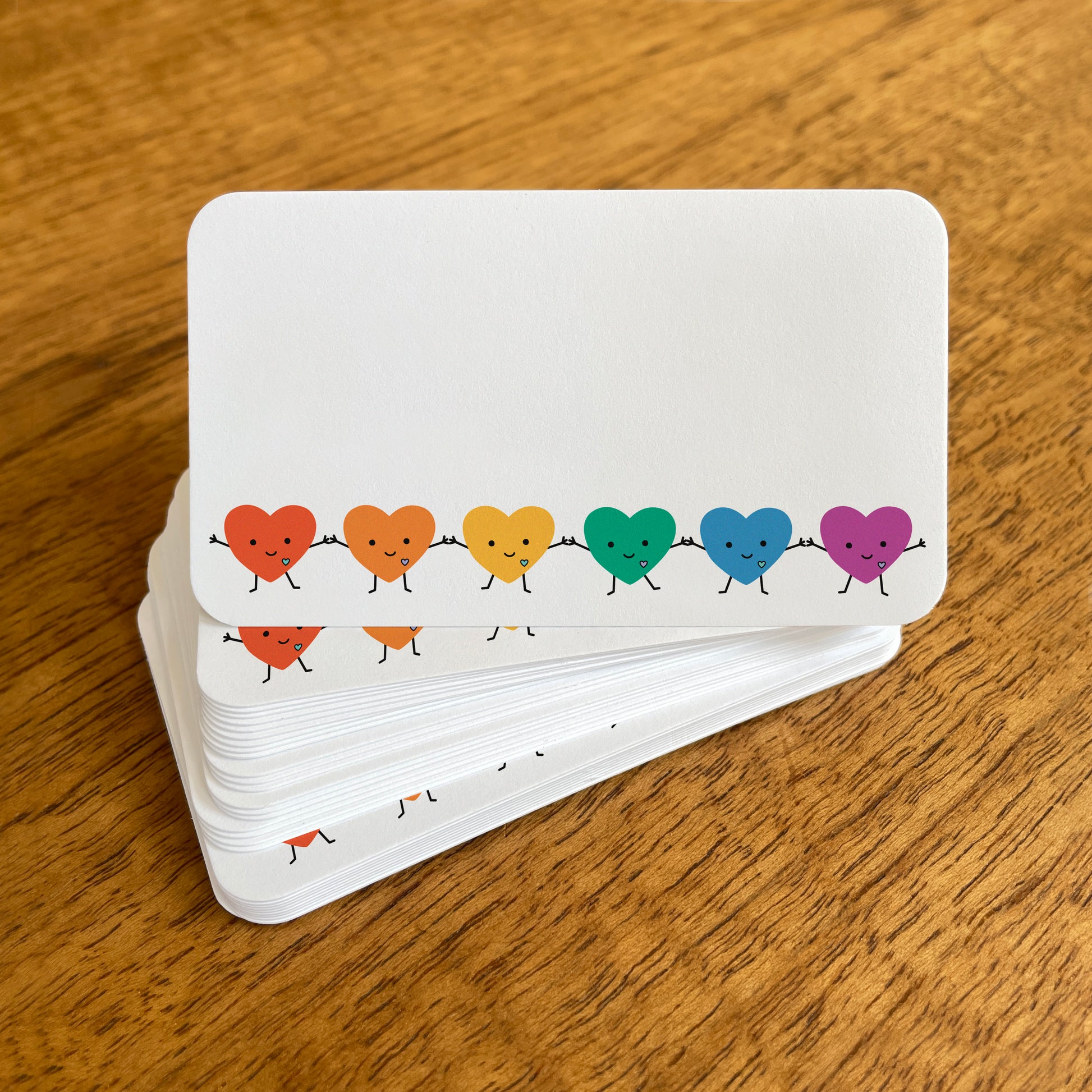 A stack of small white Happy Hearts mini note cards with rounded corners, featuring a row of smiling rainbow heart characters holding hands.