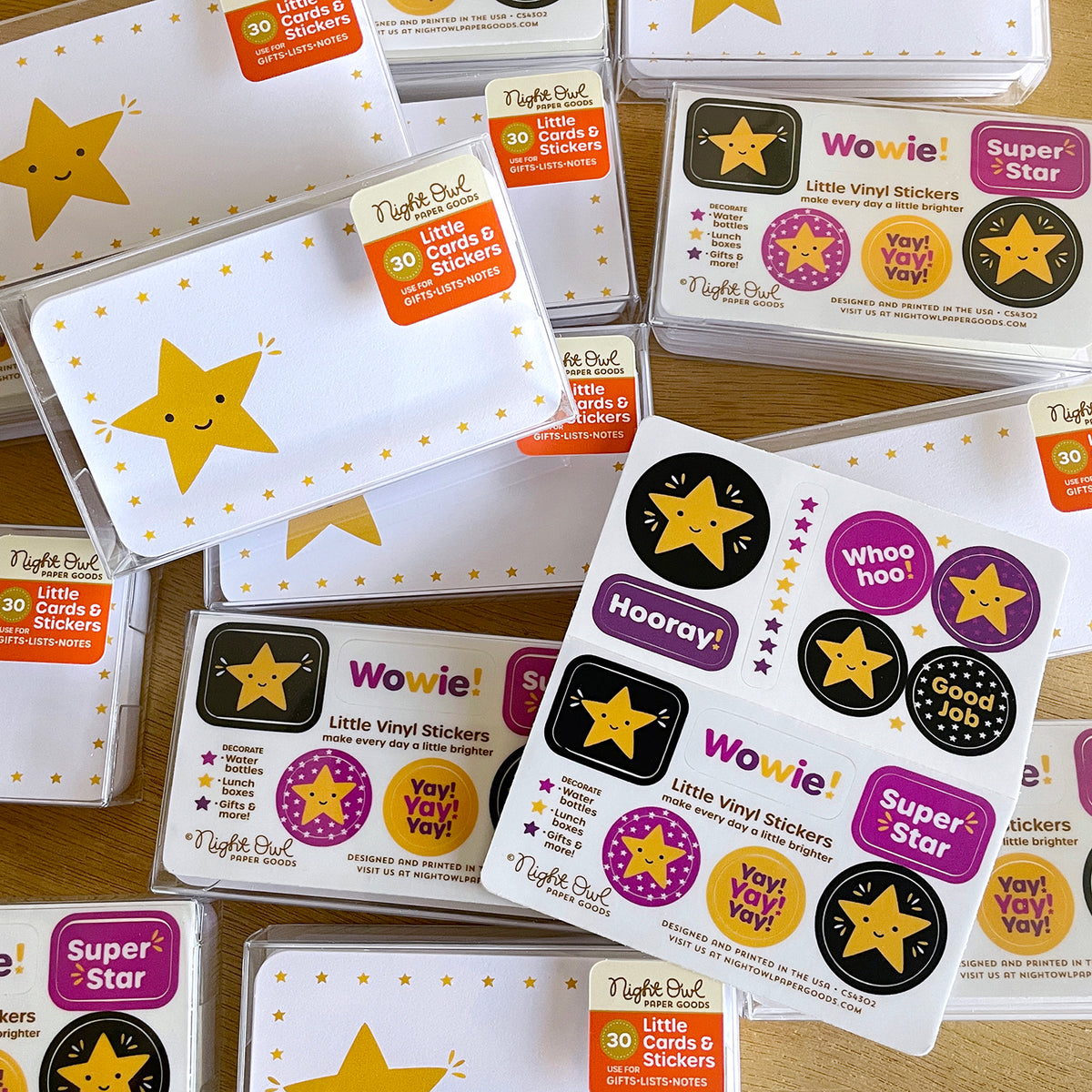 Overhead product photo of Night Owl Paper Goods Gold Star Little Cards & Stickers boxed sets, featuring smiling gold star mini note cards and coordinating colorful vinyl stickers with encouraging phrases like “Wowie!,” “Hooray!,” “Super Star” and “Good Job.” Gift-ready clear boxes are arranged on a wooden surface.