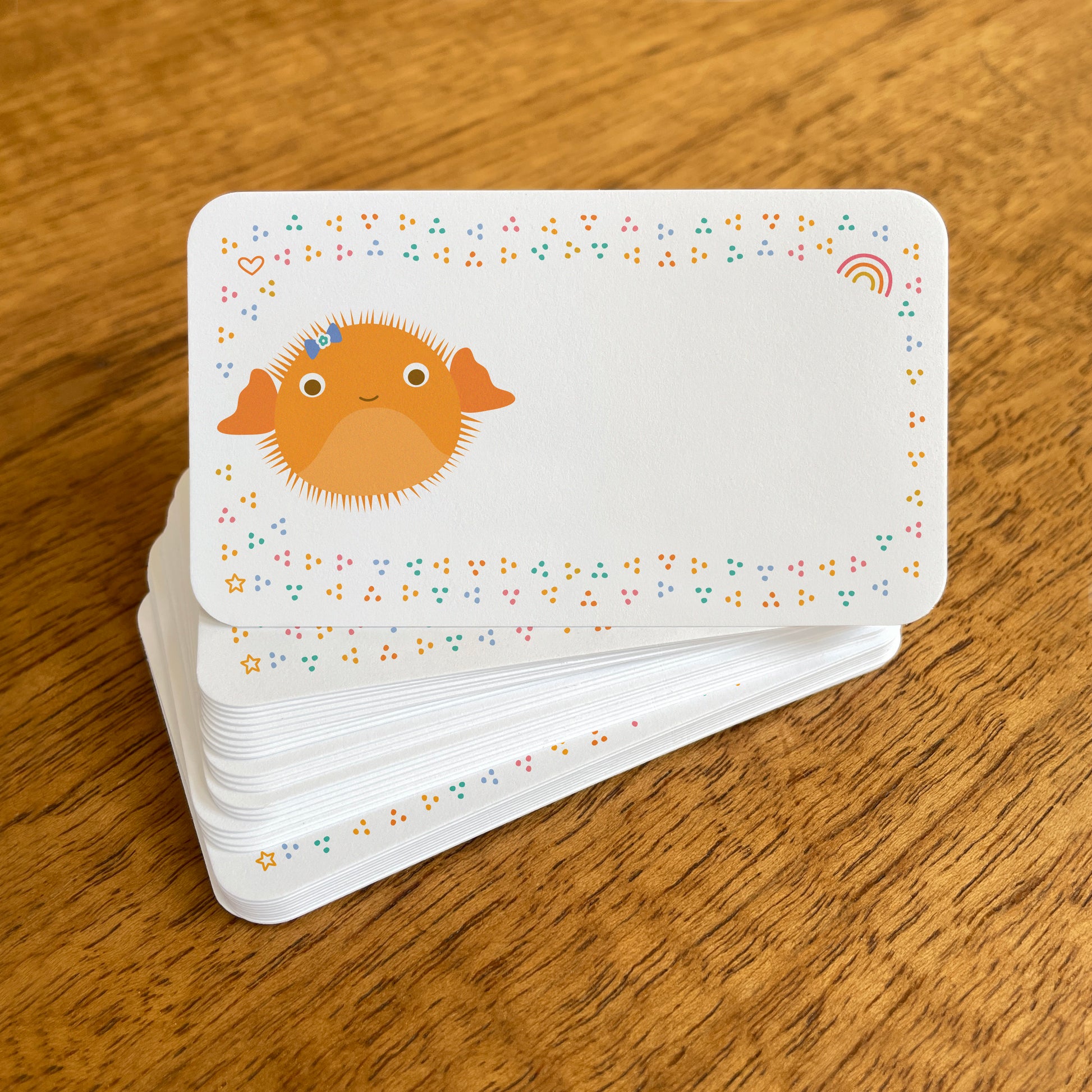 A stack of small white Puffer Fish note cards with rounded corners, featuring a cute orange puffer fish surrounded by colorful confetti-like dots, with small heart, rainbow and star details.