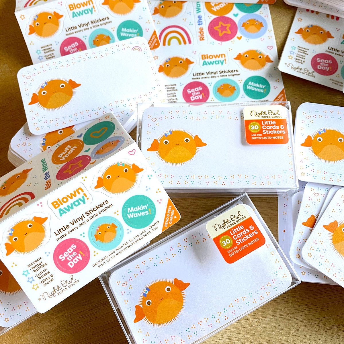 Puffer Fish Little Cards & Stickers boxed stationery sets, featuring smiling orange puffer fish mini note cards with colorful confetti accents and coordinating ocean-themed vinyl stickers with cheerful sea life illustrations and playful phrases. Several box sets, cards and stickers are arranged on a wood surface. 