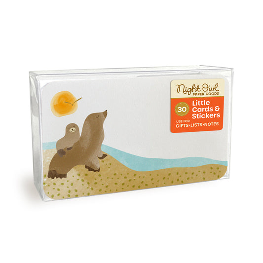 A clear plastic box displaying a small white note card with rounded corners featuring cute sea lions on a sandy beach beside the ocean. The box label reads “Night Owl Paper Goods - 30 - Little Cards & Stickers - Use for Gifts, Lists, Notes.”