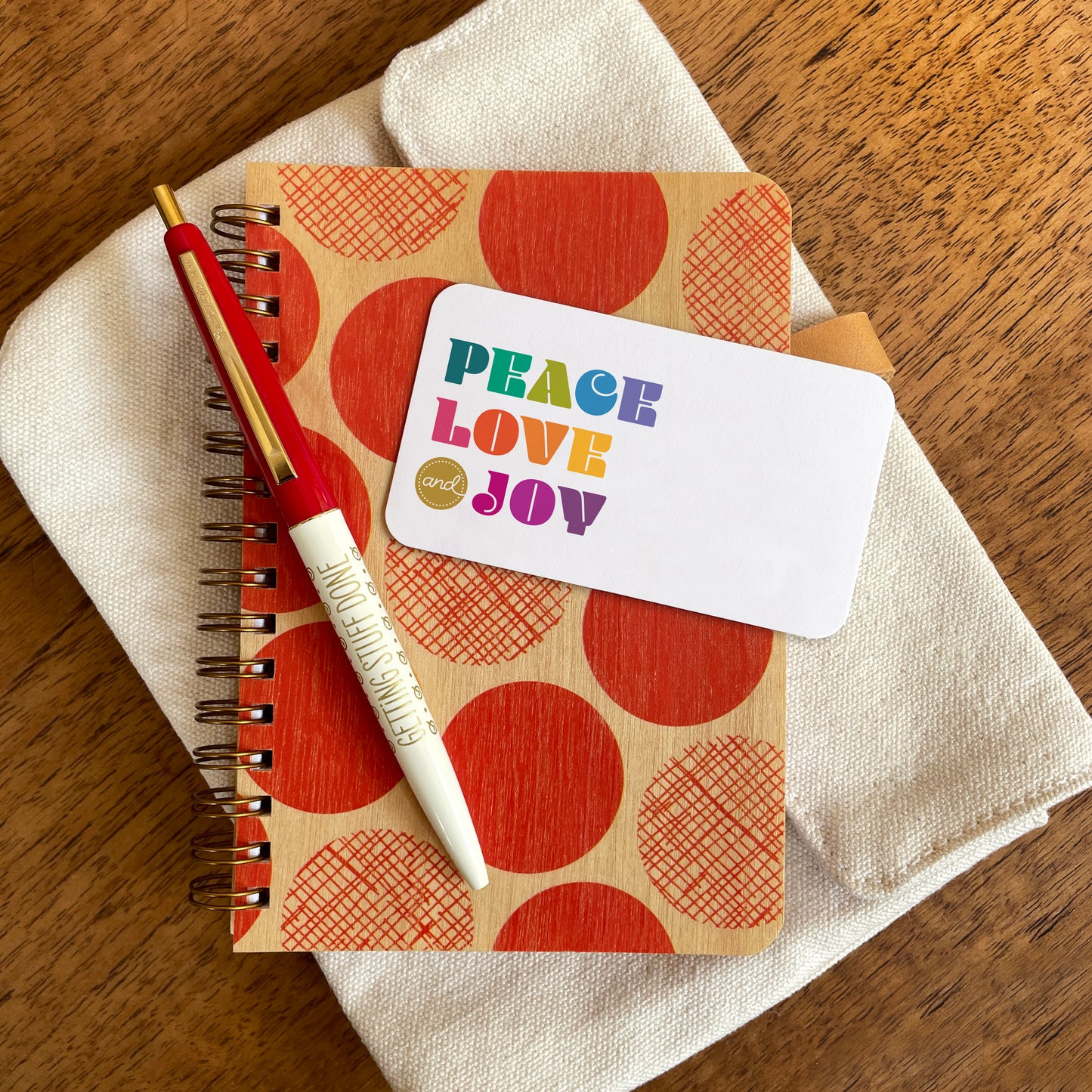 Peace Love Joy mini note card with colorful “PEACE LOVE and JOY” lettering displayed on a red polka-dot wood notebook with a red and white pen with gold trim.