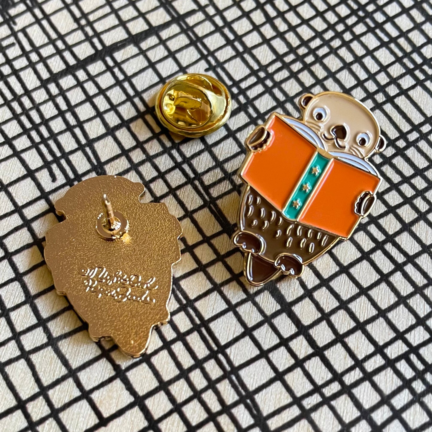 Front and back of an enamel pin of an otter reading a book. Back shows Night Owl Paper Goods script logo.