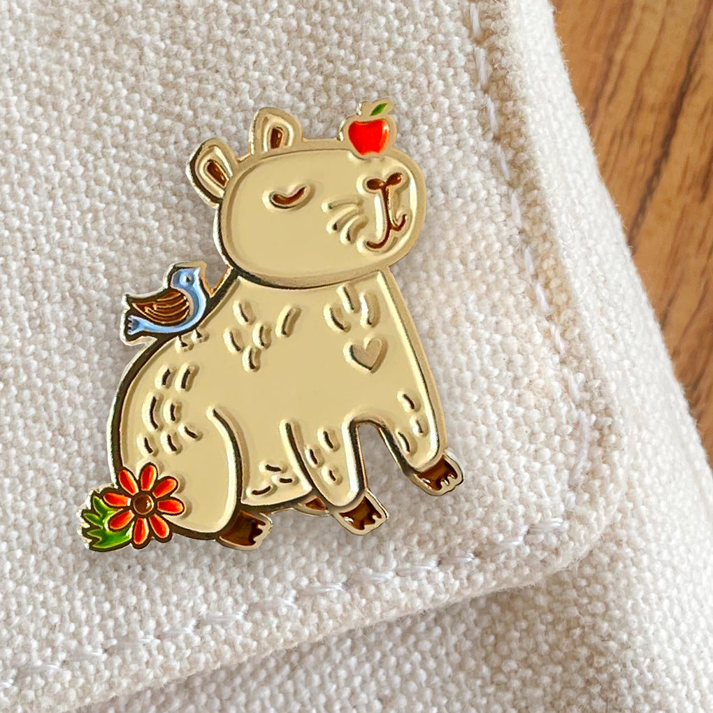 Capybara enamel pin with vibrant enamel and shiny gold metal accents featuring a smiling capybara with an apple on its head, as well as bird and flower details.