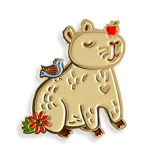 Capybara enamel pin with vibrant enamel and shiny gold metal accents featuring a smiling capybara with an apple on its head, as well as bird and flower details.