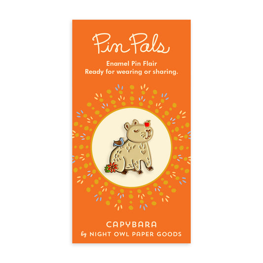 Capybara enamel pin on vibrant orange backer card. Pin features a smiling capybara with an apple on its head, as well as bird and flower details and gold accents. Backer card reads "Pin Pals Enamel Pin Flair Ready for wearing or sharing. CAPYBARA by Night Owl Paper Goods.