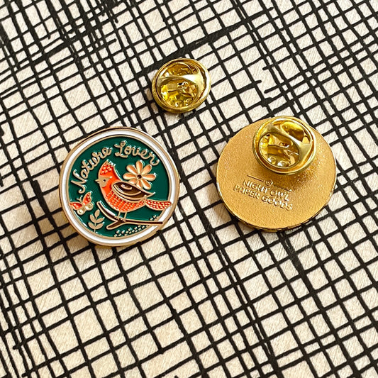 A round enamel pin shown from both front and back on a black-and-white crosshatch background. The front features a blue background with a red and brown bird, floral design and 'Nature Lover' text, outlined in gold. The back is all gold with a butterfly clasp and Night Owl Paper Goods logo.