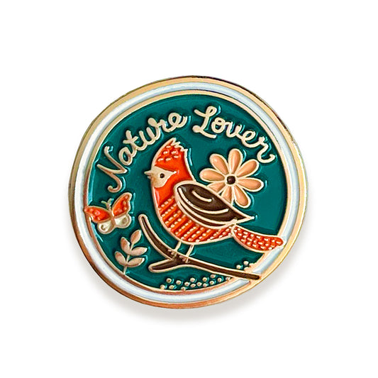 A round enamel pin featuring a blue background with the words 'Nature Lover' in white script, a red and brown bird on a branch, a peach flower and a butterfly, all outlined in gold.