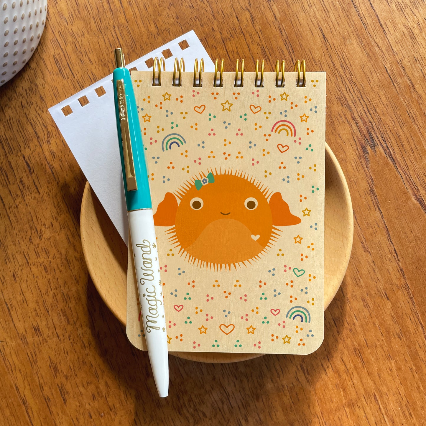 Mini notepad with birch wood covers, spiral binding at top and a cute orange puffer fish, surrounded by rainbow, star, heart and dot designs. Pictured with an aqua and white ballpoint pen that has gold accents and gold "Magic Wand" text. Both are sitting on a wood surface.