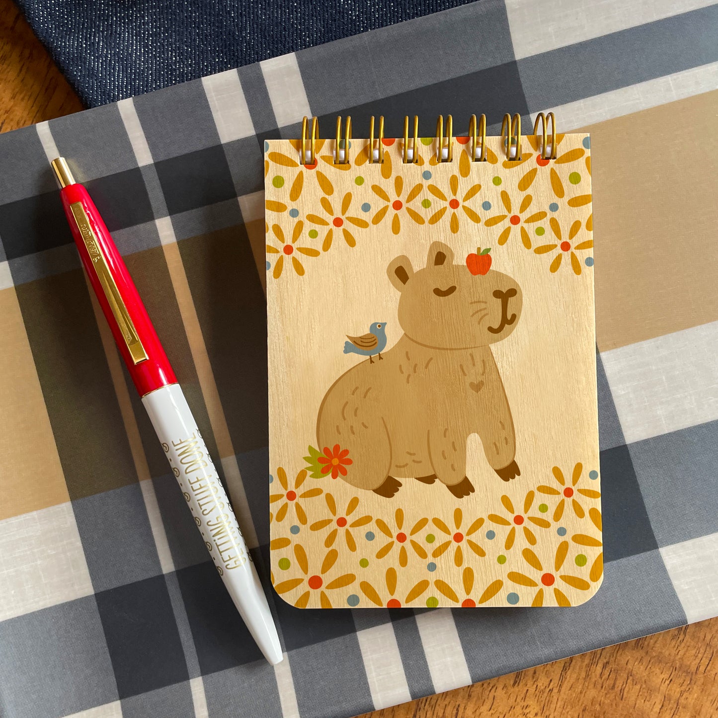 Mini notepad with birch wood covers, spiral binding at top and a content capybara with an apple on its head and a bird on its back, surrounded by floral designs. Pictured with a red and white ballpoint pen that has gold accents and gold "Getting Stuff Done" text. Both are sitting on a blue, tan and white plaid surface.