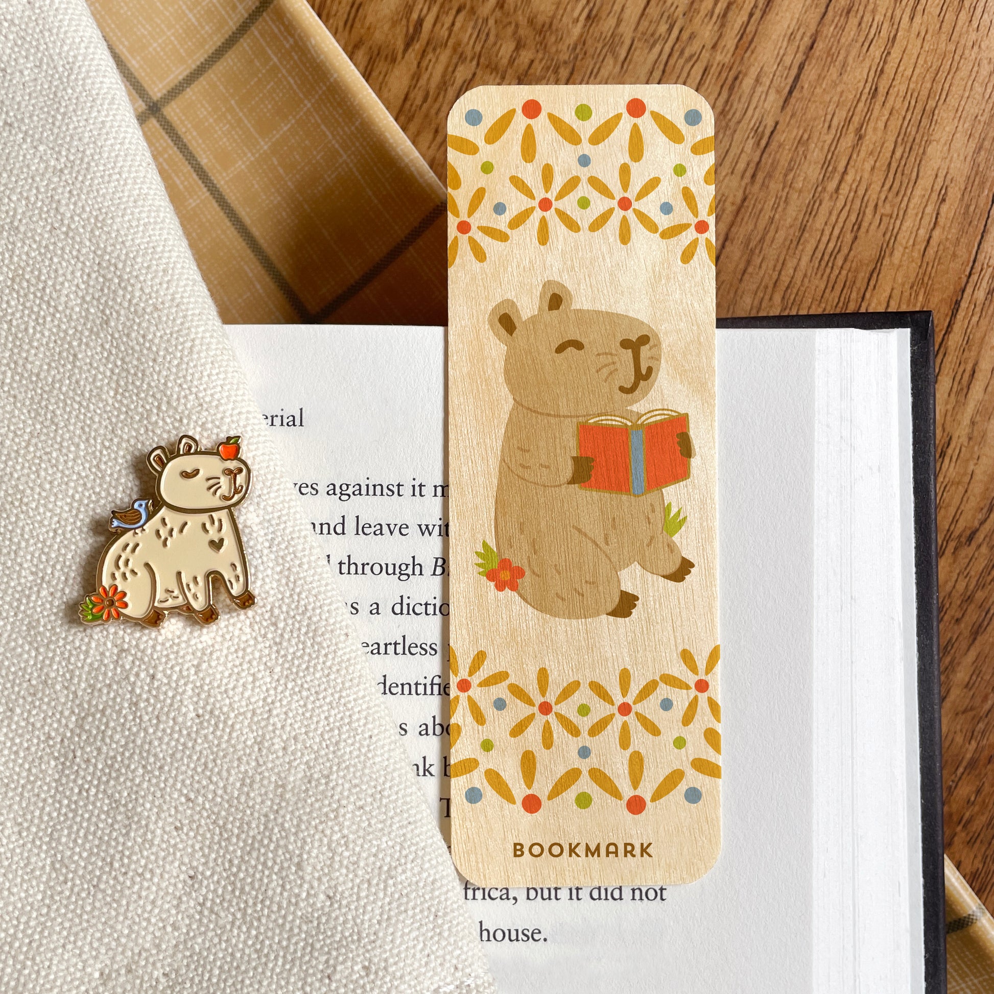 A wooden bookmark featuring a capybara reading a book, placed on an open book alongside a coordinating capybara animal pin.