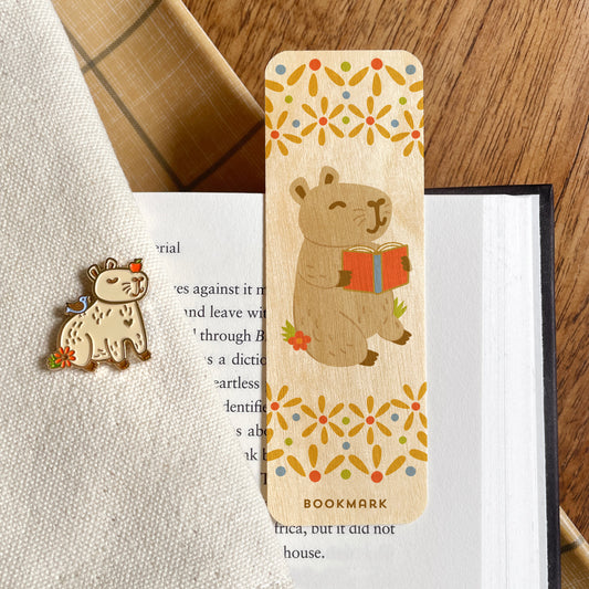 A wooden bookmark featuring a capybara reading a book, placed on an open book alongside a coordinating capybara animal pin.