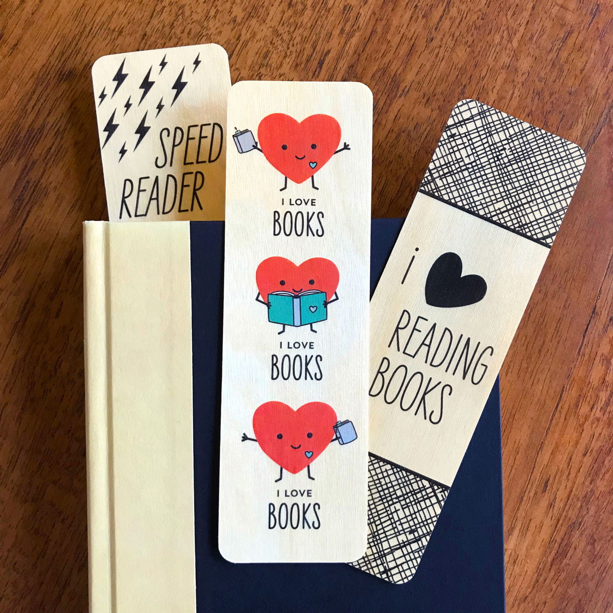 Photo of three wooden bookmarks in and on a black book. Speed Reader design has black lightning bolts, red cartoon happy hearts are reading books and black pattern with text that reads ' i heart reading books'.