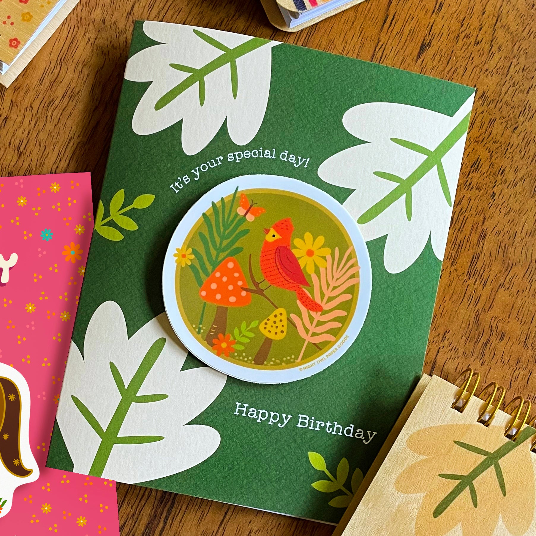 Green birthday card with a removable red bird vinyl sticker and nature-themed illustrations displayed on a wooden surface