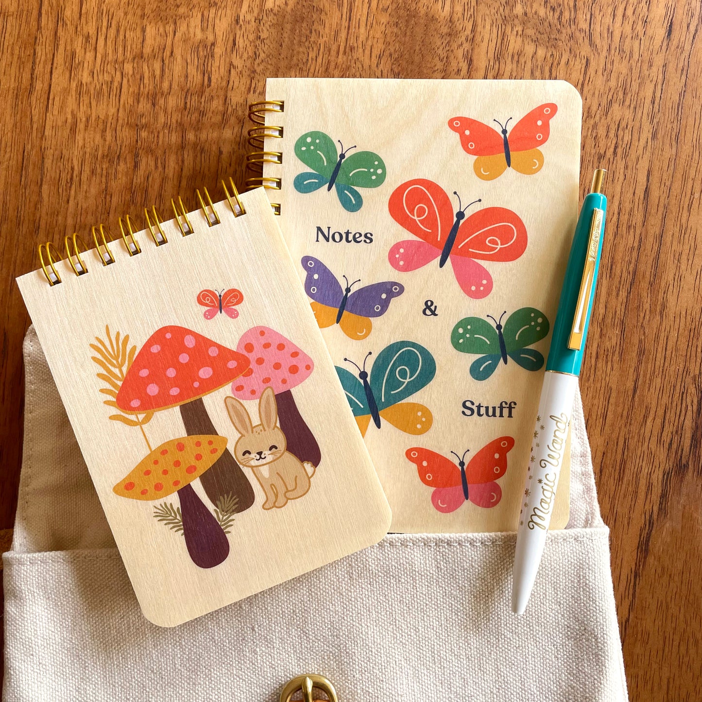 Mini wood notepad with rabbit and mushrooms alongside a pocket-sized wood notebook with butterflies and a Magic Wand ballpoint pen.