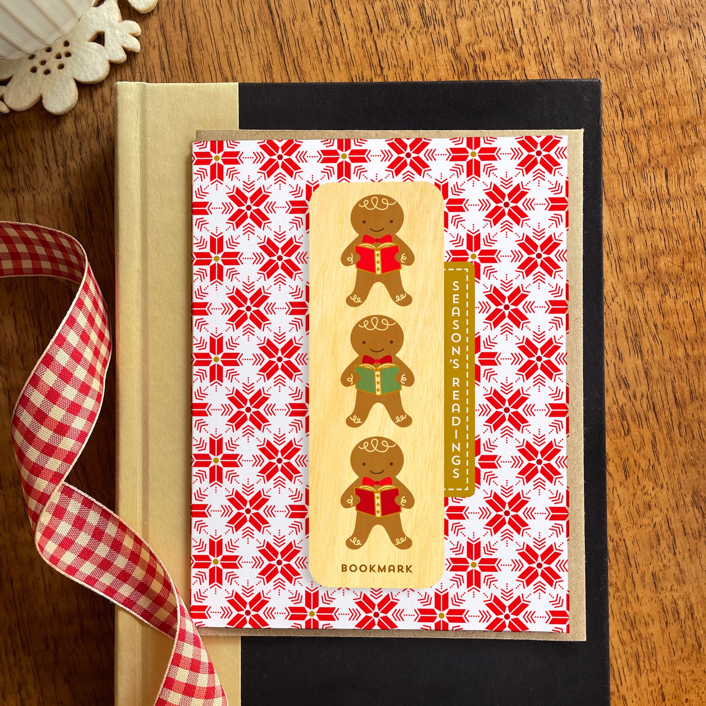 Folded holiday greeting card with red and white snowflake pattern, “Season’s Readings” text and a removable birch wood bookmark featuring three gingerbread cookies holding books. Pictured with a kraft envelope and sitting atop a book on a wooden table with a red gingham ribbon.